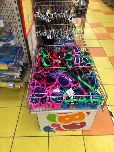 The image provides a candid view inside a retail store, likely a discount or toy shop, characterized by walls tiled in a checkerboard pattern of yellow and orange. The central focus is a large, overflowing wire basket fixed to a shelf, densely packed with numerous oversized novelty sunglasses. These sunglasses come in a vibrant palette of colors, including bright pink, neon green, red, and blue, many still bearing small white price tags.
To the left of this display, another rack holds rows of standard-sized sunglasses, most appearing to be individually packaged in clear plastic. Below the main sunglasses display, a lower shelving unit is visible, showcasing a variety of blister-packaged toys hanging on hooks. Among these, several Hot Wheels toy cars are clearly identifiable by their distinctive packaging and branding. Visible price tags on these toy packages indicate "1,50 €".
On the far right, partially obscured, is a large cardboard box or sign with colorful graphics, featuring prominent rounded letters like 'B' and 'O', suggesting part of a brand name or logo. The store's interior is illuminated by artificial lighting, and no people are present. A price tag on one of the sunglasses also shows "1,00 €". The scene captures a typical retail merchandising setup, conveying a bright, if somewhat cluttered, atmosphere in a store located in Marousi, Greece.