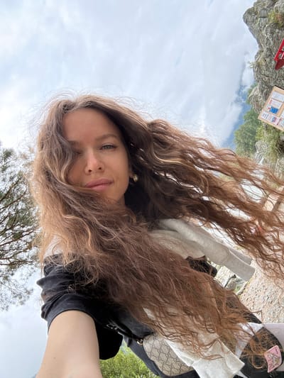 A young woman with long, wavy, light brown hair is taking a selfie outdoors. She is wearing a black t-shirt and a light grey hoodie. Her hair is partially covering her face, but her green eyes, nose, and lips are visible. She is smiling softly.
In the background, there are rocky formations, lush green trees, and a cloudy sky. There appears to be a bulletin board with some papers attached to it, and a red object that might be part of a sled or another outdoor equipment. The scene suggests an outdoor recreational area, possibly in a mountainous or hilly region.
The image is oriented sideways, with the woman's head at the bottom and the sky at the top, indicating it was taken with the phone held vertically and then rotated. The lighting suggests it is daytime, with overcast skies. The overall mood is relaxed and natural.
No visible text is legible in the image.
Location context provided: Elekmonar, Russia.