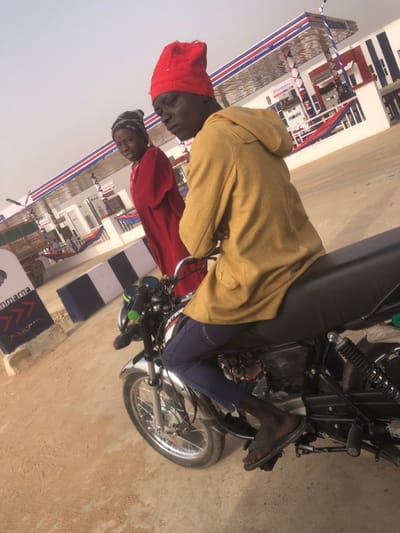 Two men stand near a motorcycle. One man wears a red head covering and red shirt, while the other wears a tan jacket. They are positioned in front of a building decorated with banners and flags in red, white, and blue. The motorcycle is black with silver wheels. The ground is light brown and appears to be a sandy or dusty surface. The sky is overcast.
