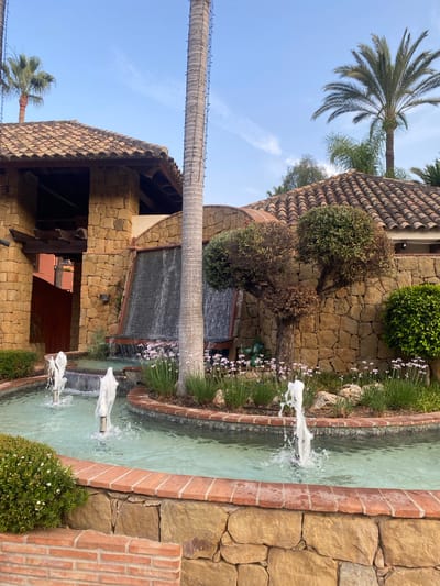 The image shows a man-made water feature with multiple fountains and a cascading waterfall. The architecture appears to be of Mediterranean or Spanish style, with stone walls and terracotta tiled roofs. Lush greenery, including palm trees and various shrubs and flowers, surrounds the water feature, suggesting a warm climate. The lighting indicates it is daytime, possibly late afternoon given the soft light. The scene is tranquil and suggests a well-maintained outdoor space, likely a resort, hotel, or private residence in a coastal town. Without additional information or context, pinpointing the exact location within Estepona, Spain, is impossible, but the architectural style and vegetation are consistent with the region. There are no people visible in the image.