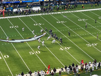 An aerial view shows an American football game in progress. Two teams line up on the field. One team wears white and brown uniforms, the other navy blue and white. They are positioned near the 40-yard line. The field is green with white yard lines and numbers. Spectators fill the stands in the background. The Chargers' logo is visible on the field. There are sidelines with referees and coaches.
