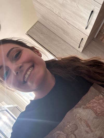 In this selfie, a young woman with brown hair and a black t-shirt smiles widely. She is positioned indoors, with kitchen cabinets visible in the background, suggesting a domestic setting. A window with textured glass and sunlight streaming through is also visible. The image was taken from a low angle, looking up at the woman. The location is identified as Bulgarovo, Bulgaria.