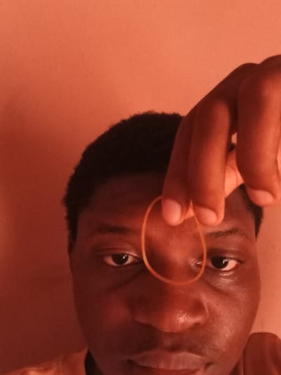 A close-up, overhead shot shows a person's face in Bauchi, Nigeria. The person's skin tone is dark, and they have short, dark hair. They are looking directly at the camera. A rubber band is held in front of their face, obscuring part of their nose. The background is a solid, orange hue.