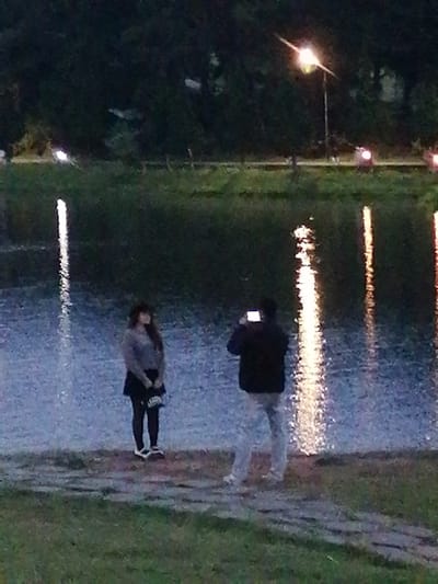 The image depicts two individuals near a body of water, possibly Xuan Huong Lake in Da Lat, Vietnam, during twilight. On the left, a woman stands facing slightly away from the camera, wearing a dark skirt, black tights, and white shoes, with a dark jacket or sweater and a dark hat. She is holding a dark object. To her right, a man is positioned in profile, facing the woman and holding a smartphone in his hands, appearing to take a photograph of her. He is wearing a dark jacket and light-colored pants. The water's surface is illuminated by reflections of lights, likely streetlights and perhaps the moon, creating bright vertical streaks. The background consists of a dark, tree-lined shoreline and a street with a lamppost visible in the upper right, emitting a bright light. The overall scene suggests a calm, possibly romantic or casual, moment by the lake.