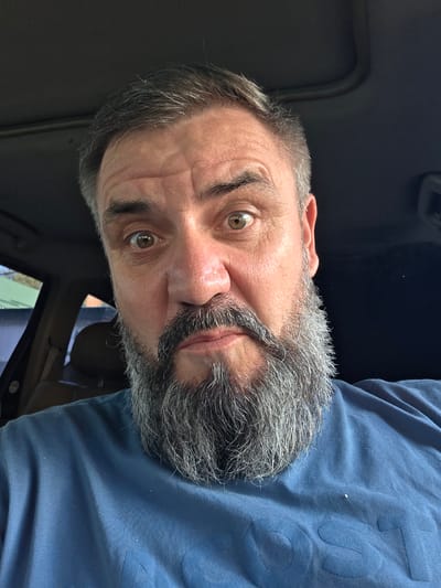 A man with a full grey beard and side-swept dark hair is shown in a close-up, sideways selfie. He is wearing a blue t-shirt, and his face is illuminated by natural light, suggesting it is daytime. The man's eyes are open, and he appears to be looking directly at the camera. His expression is neutral, with a slight furrow in his brow. The background is dark and appears to be the interior of a car, with hints of a sunroof and the car's interior trim. The overall scene suggests a casual, everyday moment captured by the man himself. There is no discernible text in the image.
