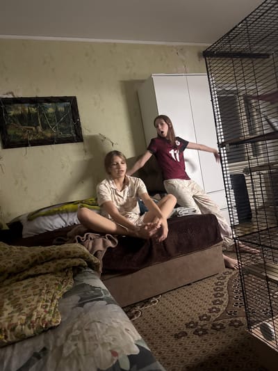The image depicts an indoor scene, likely a bedroom, with two young individuals present. One individual, on the left, is seated on a bed, wearing light-colored pajamas. They are positioned facing forward and their legs are extended. The second individual, on the right, is standing and appears to be gesturing with their arms. They are wearing a dark red t-shirt with the number 11 on it and patterned pants. To the right of the standing individual is a large black wire cage. The room's decor includes wallpaper with a textured pattern, a framed picture on the wall, and a patterned rug on the floor. The lighting suggests it is nighttime or a dimly lit environment. The image does not provide any specific information to determine its location within the city of Shakhty, Russia.
