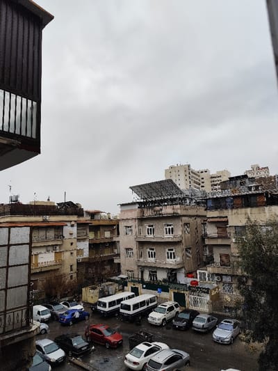 The image presents an urban landscape within As-Salihiya Municipality, Syria, under an overcast sky. The perspective is elevated, with a dark, vertically-slatted architectural element, likely a balcony or building exterior, partially framing the left foreground.
The mid-ground is dominated by a cluster of multi-story buildings exhibiting various architectural styles and states of repair. A prominent grey-toned building in the center-right features multiple windows and balconies, and its rooftop hosts a large, grid-mounted solar panel array. Other building rooftops display equipment such as water tanks, satellite dishes, and various utilities.
Below the buildings, an open-air parking area occupies the lower central portion of the image. This area contains numerous parked vehicles, including sedans of various colors (e.g., red, blue, silver, black), a white SUV, and two distinct white minibuses. A single individual is observed leaning over the front of a blue sedan in the parking area, suggesting an interaction with the vehicle. The ground surface of the parking area appears wet. Several dark-colored refuse bins are also visible within the parking zone.
To the right of the parking area, a tree with sparse foliage is present. In the distant background, taller, lighter-colored multi-story buildings are discernible against the grey, cloudy sky.