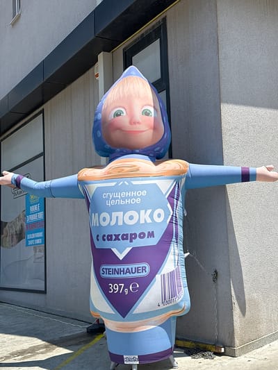 An inflatable cartoon character resembling Masha from the animated series "Masha and the Bear" stands in front of a building in Donji Pobori, Montenegro. The character is wearing a blue headscarf with white polka dots and a light blue outfit that resembles a milk carton. The carton design features Cyrillic text that reads "МОЛОКО" (milk) and "С САХАРОМ" (with sugar), as well as branding for "STEINHAUER" and a weight of "397g". The character has its arms outstretched. Behind the character is a textured grey building with a dark window frame and a black trim above it. To the left of the character, a sign with text including "PARKING JE ZA PRIMANJE ROBE" (Parking for receiving goods) is visible on the building's glass facade. The ground in front of the building is concrete, with yellow lines indicating a boundary. The scene is illuminated by natural daylight, casting shadows on the ground and building. A chain is attached to the inflatable character and secured to the building wall.