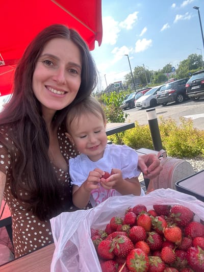 A selfie-style photograph, rotated 90 degrees clockwise, captures a woman and a young child outdoors, sharing a moment over fresh strawberries. The woman, with long, dark hair and a warm smile, is positioned in the lower-left foreground, wearing a brown top or dress with white polka dots. Her right arm is visible, and her hair flows across the frame.
Resting against the woman's shoulder/chest is a young child, also smiling with eyes closed slightly, wearing a white top. The child holds a strawberry in one hand. To the right, a large clear plastic bag overflows with ripe red strawberries, some of which are scattered on a wooden table. The woman also holds a strawberry in her left hand.
In the background, under a bright blue sky with scattered white clouds, is a parking lot or road lined with cars and green bushes. A black car with the license plate "SCL 8927H" is clearly visible, along with a blue circular traffic sign featuring a white arrow pointing right. A light-colored patterned bag, possibly a purse, rests on the table behind the strawberries. Part of a large red umbrella is visible in the lower-left corner, with faint, unreadable white text. The scene conveys a cheerful, sunny daytime setting, likely in Ustroń, Poland, with both individuals expressing happiness.