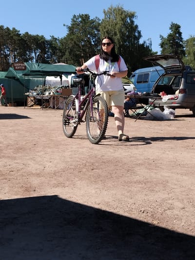 This is a full-body, eye-level, outdoor, medium shot photograph of a woman standing with a bicycle. The woman, who has dark hair and is wearing sunglasses, a white t-shirt, and light-colored shorts, is holding the handlebars of a purple bicycle. The bicycle is positioned in front of her, slightly to her right. She is wearing dark sandals. The woman is standing on a dirt surface. In the background, there are green tents and tables displaying goods, suggesting an outdoor market. Behind the tents, there are trees and a blue van with its rear door open, and a grey car. A person in a red shirt is visible on the left side of the image, walking away from the camera. The sky is clear and blue. The scene appears to be an outdoor market in Irshansk, Ukraine.