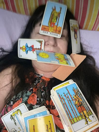 This media file captures a person lying down with several tarot cards placed on their face and body. The person has dark hair and appears to be wearing a floral-patterned shirt. The background shows a striped pillow and a colorful, patterned fabric, possibly a blanket or clothing.
Several tarot cards are visible, including:
* A card with a figure standing and holding a staff, with text that appears to be in Cyrillic script, possibly "ПАЖ ЖЕЗЛОВ" (Page of Wands).
* A card depicting the sun with a smiling face.
* An inverted card with a figure hanging upside down, also with Cyrillic text that appears to be "ПОВЕШЕННЫЙ" (The Hanged Man).
* A stack of cards, with the top one showing a figure balancing several staves, identified as card IX of Wands.
* Other cards with partial imagery of staves and Roman numerals like "III".
One of the cards is partially covered by a beige, textured object, possibly a band-aid or a similar item. The overall mood is somewhat surreal and intimate, with the cards intimately placed on the individual. The lighting suggests an indoor setting, and there are no clear indications of time of day or weather. The provided location context is Novi Petrivtsi, Ukraine.
