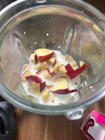 A clear glass blender jar is positioned on a wooden surface. Inside the blender jar, chopped pieces of fruit, appearing to be peaches or nectarines with red skin and yellow flesh, are submerged in a white liquid, likely milk or yogurt. The blender jar has measurement markings on its side. To the left of the blender, a portion of a black blender base is visible. In the lower right corner, a partially opened pink and white box with a red floral pattern is present. The scene is indoors, and the lighting suggests it is daytime. There are no people or animals visible in the image. The city of Gelsenkirchen, Germany is not depicted or suggested in this image.