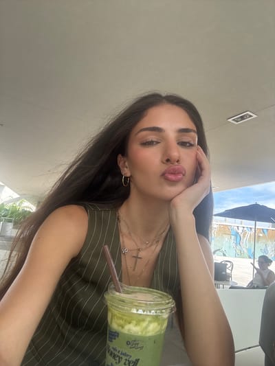A young woman with long, dark hair, wearing a green pinstripe vest and multiple necklaces, poses for a selfie with her hand on her cheek and her lips puckered. She is holding a green beverage in a clear plastic cup with a straw. In the background, there is a wall with colorful graffiti, an umbrella, and other patrons sitting at tables. The scene appears to be an outdoor seating area of a cafe or restaurant in Hallandale Beach, Florida, USA, under a covered patio with a visible sky.