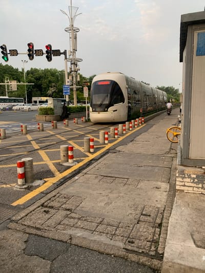 The image shows a modern light rail train in Wuhan, China, at the intersection of a major road and a dedicated light rail track. The light rail train is stopped at a station, its doors are closed, and no passengers are visible. It is a modern, clean-looking train with large windows and a streamlined design.
There are a row of red-and-white bollards along the side of the light rail track, forming a barrier to keep pedestrians and vehicles away from the tracks. The pavement in the foreground is cracked and worn, suggesting heavy use. The road is marked with yellow lines and a series of crosswalks. Traffic signals with the numbers 88 are visible above the road. To the right of the frame, a section of a white-painted wall is visible, part of an adjacent building. A single bicycle leans against the wall. The scene is typical of a bustling urban environment, with a mixture of modern and older infrastructure, suggesting a city in a state of rapid growth and development. The coordinates provided place the scene in the eastern part of Wuhan.