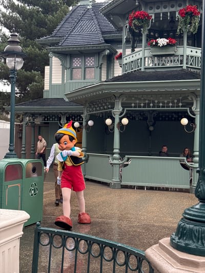 The image captures an outdoor scene at Disneyland Paris in Chessy, France, under overcast conditions with visible moisture on the pavement. A person in a full Pinocchio character costume stands prominently in the midground, wearing a yellow shirt, red shorts, black vest with gold trim, blue hat with a red feather, white gloves, and large brown shoes. The character's hands are positioned near its chest. Behind Pinocchio, a male handler in a light-colored vest, white shirt, and dark trousers is partially visible walking towards the left.
The background features an ornate, multi-story Victorian-style building painted in shades of mint green, dark green, and white. It has multiple dark, possibly slate, roofs and a distinctive turret. An upper-level open-air balcony or gazebo section of the building is adorned with red and white flowering plants in hanging baskets and window boxes, enclosed by intricate white railings. The lower level features a covered walkway with numerous decorative supports and several round white globe light fixtures. Two individuals, one male and one female, are seated within this covered structure on the right side of the image.
On the far left, two dark green recycling bins with silver tops and recycling symbols are visible next to an ornate dark green lamppost with a clear globe light. Another similar lamppost base is on the far right. A dark green metal railing with circular patterns occupies the immediate foreground. The pavement is a dark, speckled material that appears wet and reflective.