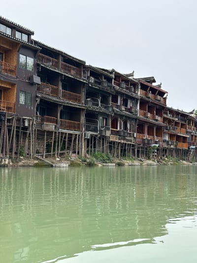 A tall, weathered building, reminiscent of traditional Chinese architecture, stands alongside a body of water. The building features multiple levels with exposed wooden frameworks, balconies, and visible interior spaces. Its facade is aged and appears to be in a state of partial disrepair. The water's surface reflects a distorted image of the building, creating a mirror effect. The environment suggests a location in Fenghuang, China, where such structures are common along the Tuojiang River. The sky above is overcast, casting a muted light on the scene. There are no people visible in the image.