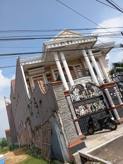 The image shows a two-story house with a white facade and ornate details, situated in Kutawaringin, Indonesia, at coordinates -6.978, 107.549. The house has a balcony with a wrought iron railing and several large columns. The house is partially obscured by a tall, gray wall with a wooden fence and a gate with a black wrought iron design. A black motorcycle is parked on the ground in front of the gate. There are numerous electrical wires running across the sky, and a clear blue sky above. The image is taken from a low angle, looking up at the house and the wall.