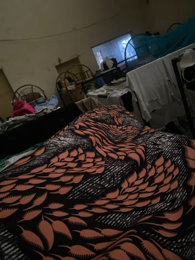 This is a close-up, low-angle shot of a person lying on a bed covered with a patterned orange and black fabric. The fabric has a repeating motif of leaf-like shapes with serrated edges. In the background, the scene appears to be a bedroom in a dimly lit room. There are multiple beds visible, some with metal frames. Various items of clothing are draped over furniture and beds, including a pink garment and a white bra with black trim. A blue mosquito net is visible over one of the beds. A window with vertical blinds is partially visible in the background. The overall environment suggests a shared living space, possibly a dormitory or a room in a multi-occupant dwelling. This scene is located in the city of Jimeta, Nigeria.