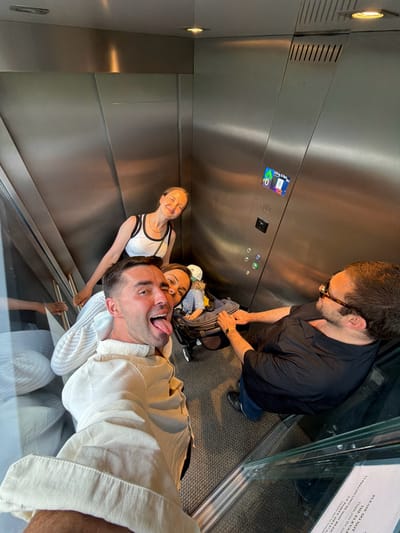 In an elevator, four individuals are captured in a selfie. The person closest to the camera is a man wearing a cream-colored shirt, sticking his tongue out and winking. To his left and slightly behind him, a woman in a white tank top with black trim smiles. Further back, a woman with short brown hair is seen, and next to her, a baby in a stroller. To the right, a man with dark hair and sunglasses, wearing a black shirt and blue jeans, stands. The elevator has metallic walls and a grey carpeted floor. A sign with Italian text is visible on the wall. The overall scene suggests a casual, possibly tourist-related moment in Pognana Lario, Italy, given the location's context.