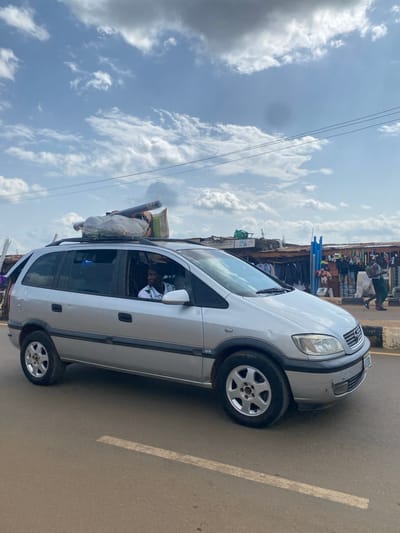 A silver minivan is parked on a paved road in Kafanchan B, Nigeria, with its roof rack loaded with various items, including bags of goods and what appears to be a rolled-up carpet or mat. A man wearing a white shirt with green trim is visible in the driver's seat. In the background, there are open-air market stalls with merchandise displayed, including clothing and other goods. The sky is partly cloudy with a mix of white and gray clouds. The scene appears to be a typical roadside market environment in a Nigerian town.