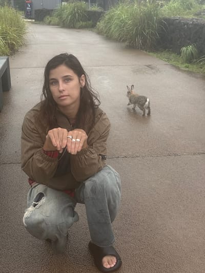 The image features a young woman with long, dark hair and striking blue eyes, crouching on a paved surface. She is wearing a brown jacket over a red and black plaid shirt, and loose-fitting denim jeans. Her hands are clasped in front of her, adorned with several rings. To her left, a small, furry rabbit is captured mid-hop on the same paved ground. Lush green vegetation lines the left side of the frame, suggesting an outdoor setting. The pavement appears damp, hinting at recent rain or mist, and the overall lighting is diffused, indicating an overcast day. The woman's expression is calm and observant, her gaze directed towards the viewer. Beside her, a grey, ripped hooded sweatshirt rests on the ground. The scene is set in Seogwipo-si, South Korea. There is some faint text visible on a sign in the background, but it is illegible.