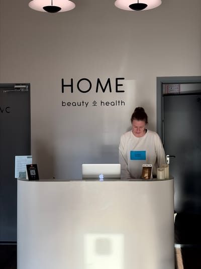 The image shows an indoor reception area, possibly in the city of Бараўлянскі сельскі Савет, Belarus, judging by the presence of Russian language text on signage. A woman with her hair in a bun stands behind a white reception desk, looking down. She is wearing a white sweatshirt with a blue rectangular patch on it. On the desk, there is a laptop with an Apple logo, a small digital display showing a QR code, and two decorative candles. To the left of the desk, there is a door with "VC" written on it, and a sign with text and a QR code. A larger sign with black lettering reads "HOME beauty & health". To the right of the woman is a dark door. The overall impression is a clean, modern reception area.