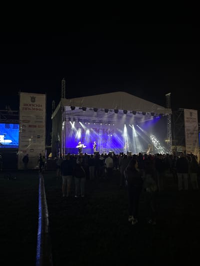 This image depicts a nighttime outdoor concert or festival in Elin Pelin, Bulgaria. A large stage, covered by a white canopy, is centrally positioned and brightly illuminated by multiple blue and white spotlights. On the stage, at least two performers are visible: one appears to be playing a keyboard or synthesizer, and another is holding a guitar. A drum kit is also present. The stage backdrop features text in Cyrillic, prominently displaying "ШОПСКИ ПРАЗНИК" (Shopska Festival/Holiday), along with a distinct, illuminated graphic design on the right side.

A sizable crowd of attendees is gathered on a grassy field in front of the stage, with most individuals facing the performers. Their forms are largely silhouetted against the stage lighting.

Flanking the stage on both sides are tall vertical banners. The banner on the left, more clearly visible, displays "ШОПСКИ ПРАЗНИК" at the top alongside a municipal coat of arms, followed by rows of sponsor logos including "ECO PETROL," "FLYNET," "dm," "MARKET," "NOBEL," "AEC-X," and "Semmelrock." To the left of this banner, a large vertical LED screen displays blue and white digital content. A person wearing a high-visibility vest stands near the base of the left banner. A bicycle wheel rests on the ground nearby.

Structural scaffolding supports the stage and banners. In the foreground, a long, narrow, luminous strip, possibly a cable protector or temporary pathway, extends across the dark grassy ground, emitting a blueish glow in sections. The overall environment outside the immediate stage area is dark, emphasizing the artificial illumination.