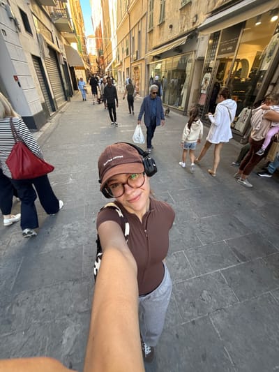 The image captures a selfie of a person with a brown cap and glasses, wearing a brown top, walking down a street in Sanremo, Italy. The pedestrian street is lined with buildings, including shops with awnings and display windows. There are other pedestrians, including individuals, families, and groups of people, walking in both directions on the cobblestone street. The lighting indicates it is daytime. One person is wearing black and white stripes and a red bag, positioned toward the bottom of the frame.