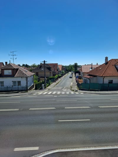 The image depicts a street scene in Bratislava, Slovakia, under a clear, bright blue sky, suggesting it's a sunny day. The perspective is high, looking down the street from a slightly elevated position. On the left side of the street, there is a row of residential houses with red tiled roofs and white or light-colored walls. Some houses have green fences and manicured hedges. Further down the street, trees and foliage are visible, indicating a suburban or semi-urban environment.

On the right side of the image is a paved road with double white lines marking lanes. A pedestrian crossing is visible near the bottom right of the frame. The road appears to be moderately busy, with several cars parked along the street and a few vehicles driving in the distance.

Traffic signs are visible on the left side of the road, including a circular sign with a red border and a diagonal red bar, likely indicating a prohibition. Another sign appears to be a blue directional sign. There are also what seem to be television antennas or radio aerials on top of some houses.

The overall impression is a calm, everyday scene in a residential area during daylight. There are no people clearly visible in the image. The lighting and clear sky suggest the time of day is likely mid-morning or afternoon.