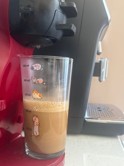 A close-up shot focuses on a clear glass filled with a frothy, light brown beverage, likely coffee, sitting on a white speckled countertop. The glass is adorned with cartoonish cat illustrations at various measurement markings: a cat with a blue and pink striped sweater holding a small object near the 500 ml line, an orange and blue cat resting near the 400 ml line, and an orange and white cat sitting near the 300 ml line. The beverage level is slightly above the 200 ml mark. A pink heart is visible near the 200 ml line.

To the left of the glass, a red coffee machine is partially visible, with its water reservoir and drip tray area in view. A single drop of liquid is suspended mid-air from the coffee machine's dispensing spout, indicating a recent or ongoing brewing process.

To the right of the glass, a black coffee machine is seen, featuring a silver drip tray with a patterned surface and a metallic trim. The machines are positioned side-by-side. The background is a plain, light-colored wall. The image composition is centered on the coffee glass and the machines, with the countertop serving as the immediate foreground. The scene does not contain any identifiable features that would place it specifically in Belashtitsa, Bulgaria; it depicts a generic domestic or office kitchen setting. There are no people or animals present in the image.