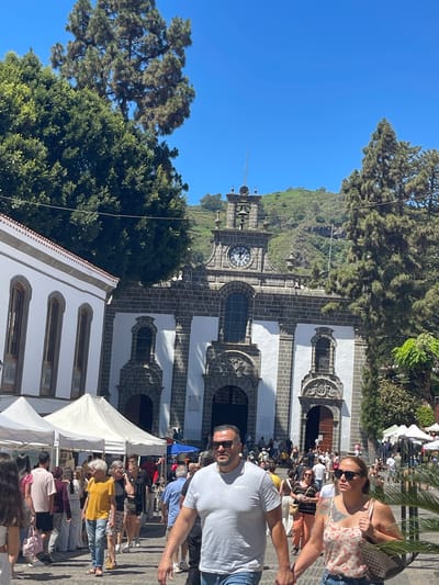 The image captures a bustling scene in the city of Teror, Spain, likely during a market day, in front of the neoclassical Basilica of Our Lady of the Pine. A crowd of people are walking along a cobblestone street, with several white market stalls set up on the left side of the image. The basilica, constructed with dark stone and white plaster, stands prominently in the background with its clock tower and bell tower visible. Lush green trees frame the scene, and a hilly, verdant landscape rises behind the basilica. The sky is a clear, bright blue, indicating a sunny day. The individuals in the foreground are casually dressed, suggesting a relaxed, everyday atmosphere. A couple holding hands is in the immediate foreground, with other pedestrians moving through the square. The overall impression is of a vibrant community gathering in a historic and scenic location.