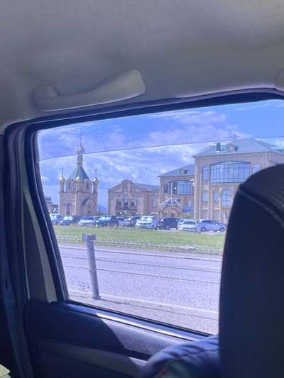 The image, taken from inside a vehicle, captures a view through the side window of buildings and a sky filled with white clouds. In the background, a distinctive religious building with multiple golden domes and a green roof stands prominently. Adjacent to it, another building with a light-colored facade and arched windows is visible. A line of cars is parked in front of these structures. The foreground shows the interior of the car, including a dark-colored seat and part of the window frame. The environment appears to be a sunny day. The text on the window provides some details about the car's certification and manufacturer.