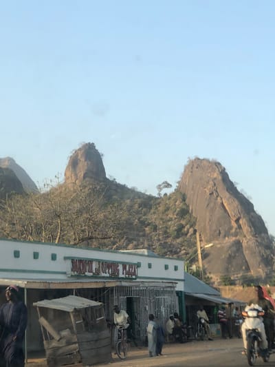 The image depicts a street scene in Bununu East, Nigeria, characterized by prominent rocky hills under a clear, light blue sky. In the foreground, a single-story building identified as "BANDIRI SHOPPING PLAZA" occupies the lower mid-section. The building is white with a light green/teal trim along its roofline and features multiple entrances, at least one secured by a metal gate.

In front of the building, a rustic, unpaved road or dirt area is visible. A small, weathered wooden kiosk or stall stands on the left. Approximately 8-10 individuals are present on the street or near the building. On the far left, a person in dark blue clothing is partially visible, facing away. A man rides a red bicycle towards the right in the mid-ground. Several other individuals, primarily men dressed in traditional attire, are observed standing or seated along the front of the plaza. On the far right, a white motorcycle with two riders is partially visible, moving right. A utility pole stands upright on the right side, against the backdrop of the hills.

The background is dominated by two large, rounded granite inselbergs or rock formations, characteristic of the Nigerian landscape. These hills are sparsely covered with dry, deciduous-looking trees and shrubs, indicating a dry season or arid environment. The sky above is clear and uniformly light blue.