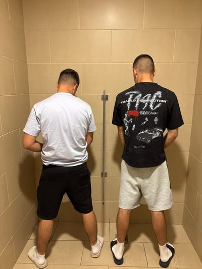 Two men are standing at urinals in a public restroom. The restroom has tile walls and floors. One man is wearing a black t-shirt with white graphics on the back, gray shorts, and white socks with black slides. The other man is wearing a white t-shirt, black shorts, and white sneakers with socks. Both men are facing the urinals and appear to be urinating. There is a metal bar mounted on the wall between the two urinals.
Kalotina, Bulgaria, is not discernible from the image.