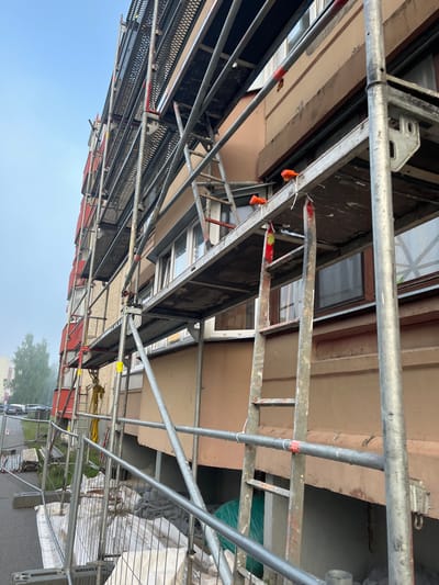 The image depicts scaffolding erected on the exterior of a multi-story building, likely an apartment block, in Maardu, Estonia. The building's facade appears to be undergoing renovation or repair, as indicated by the extensive scaffolding and visible building materials. The scaffolding is constructed from metal poles and platforms, with some sections covered in mesh. Safety barriers are installed at ground level, enclosing the construction area. The scene is somewhat obscured by fog or mist, creating a hazy atmosphere, particularly in the background where trees and other buildings are faintly visible. Several white tarps or bags are piled near the base of the building, suggesting stored materials or debris. No individuals are clearly visible in the frame. The overall impression is one of ongoing construction work in a residential urban environment.