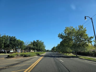 The image depicts a clear blue sky over a suburban street in Elk Grove, California. The perspective is from a moving vehicle, looking down a two-lane road lined with trees. On the left side of the road, there is a landscaped median with a "Laguna Reserve" sign and a "Safeway" store visible in the background. The road has yellow double lines indicating traffic separation and white lane markers. A street lamp is visible on the right side of the image, with power lines stretching across the sky. The trees are lush and green, suggesting a temperate climate. No people are clearly visible in the image. The overall environment is peaceful and well-maintained, typical of a residential or commercial area.