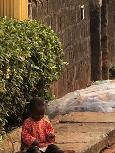 The scene depicts an outdoor area in Neni, Nigeria. A single young child, appearing to be a toddler with dark skin and short dark hair, is seated on a rough grey concrete ledge. The child wears a long-sleeved orange shirt with white polka dots, dark bottoms, and a white undershirt, and is observed looking down at a small object held in their hands. To the child's left and rear is a dense green shrub. Behind the child and to the right, a dark, textured wall, likely concrete or brick, rises with vertical striations, bearing a white numeral "1" in its upper right section. Adjacent to the child, on the concrete platform, a quantity of clear plastic bottles, some containing colored liquids, is partially obscured by a translucent white plastic covering. A vertical yellow structure is partially visible in the upper left corner.