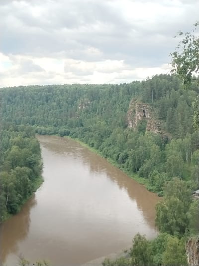 The image captures a scenic view of the Usva River as it flows through a forested landscape near Idrisovo, Russia. The river is wide and murky, with a light brown hue, indicating potentially high water or sediment content. It winds its way through a dense forest that covers the rolling hills on either side. The vegetation is predominantly deciduous trees with lush green foliage, creating a thick canopy. On the right bank, a prominent rock formation rises from the forest, its weathered surface showing signs of erosion and a dark opening that could be a cave or crevice. The sky is overcast with a layer of grey clouds, suggesting a mild or possibly rainy day. A small, rustic wooden building with a dark roof is partially visible nestled amongst the trees on the right bank, near the river's edge. The overall impression is one of a remote and natural environment.