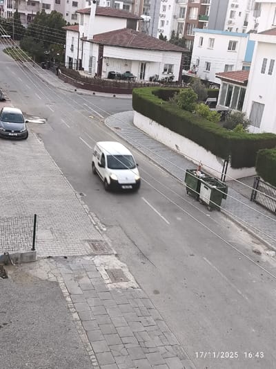 The image depicts a residential street scene in Nicosia, Cyprus, captured on 17/11/2025 at 16:43. The perspective is elevated, looking down an asphalt road that curves slightly to the right in the distance.
The road features a solid white center line and multiple overhead thin white lines, possibly utility lines, traversing the frame horizontally. Paved sidewalks with grey rectangular pavers line both sides of the street. On the left sidewalk, a dark grey Volkswagen Golf (license plate "CN 092") is parked, facing away from the camera. In the right lane of the road, a white commercial van, blurred due to motion, travels away from the viewer.
To the left and in the background, several multi-story modern apartment buildings with white facades are visible. In the midground, on the right side of the street, stands a detached white house with a reddish-brown tiled roof and dark trim, featuring a patio area with visible outdoor furniture. Adjacent to this house, along the curve of the road, a low white wall is topped by a dense, trimmed green hedge. Beyond the hedge, a portion of another white residential building is visible.
Two large, green communal waste bins on wheels are positioned on the right-hand sidewalk, near the hedge. In the bottom-left foreground, a small section of a black metal fence is present. No individuals are visible within the scene. The lighting suggests late afternoon daylight conditions.