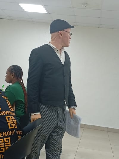 The image captures an indoor scene, likely a meeting room, classroom, or office environment. The central figure is a person with albinism, characterized by light skin and possibly light hair (though covered by a hat). This individual is a man, seen from the waist up, facing to his right (viewer's right) and looking off-camera. He is wearing a dark, possibly black, flat cap, eyeglasses, and a light-colored, collared shirt (appearing white or off-white) under a dark, possibly black, cardigan or vest. His trousers are distinctively striped with vertical grey and black lines. A metallic watch is visible on his left wrist. In his right hand, he holds a folded piece of light-colored material, possibly paper or a thin cloth.
To the left of the main subject, another person is partially visible. This individual is wearing a green t-shirt and has dark, braided hair. In the immediate foreground on the left, the back and left shoulder of a third person are visible, wearing a black top adorned with a prominent yellow or gold "GIVENCHY" logo repeated in a geometric pattern. A black chair is also partially visible in the foreground.
The room has white or very light-colored walls and a ceiling with square acoustic tiles and recessed fluorescent light fixtures providing bright illumination. A small, circular smoke detector or similar device is mounted on the ceiling in the upper right portion of the image. The floor consists of light-colored tiles. The overall impression is a well-lit, functional indoor space. The activity appears to be a gathering or a movement within such a space, with the main subject possibly addressing someone or observing something in the direction he is looking.