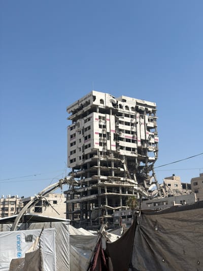 The image shows the heavily damaged exterior of a multi-story building in the city of Gaza, Palestinian Territories. The building's facade is severely compromised, with many floors collapsed and sections of the structure missing, revealing the internal skeletal framework of concrete and rebar. The exterior walls are stained and broken, with visible damage to windows and balconies. Some sections of the building are rendered to be white, with intermittent pink horizontal bands visible on some floors. The top of the building has ornate architectural details, but these are also partially destroyed.

In the foreground, there are makeshift shelters constructed from tarps and white fabric. One of the white tarps has the blue UNICEF logo prominently displayed, indicating humanitarian aid presence. The ground appears to be a mixture of debris and dirt. Electrical wires are strung across the scene, some of which are sagging or broken. Other damaged buildings and structures are visible in the background, suggesting widespread destruction in the area. The sky is clear and blue, contrasting with the devastation on the ground. There are no people visible in the image.