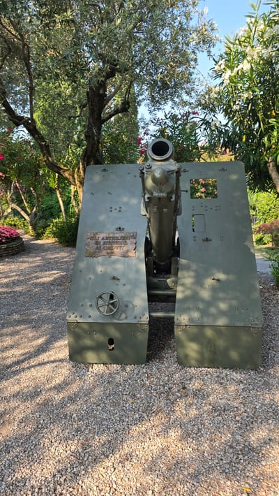 The image shows a historical artillery piece situated on a gravel path in a garden setting. The cannon is painted olive green and is prominently displayed in the center of the frame. It appears to be a static display, possibly a monument or part of a museum exhibit.

On the side of the cannon's carriage, there is a plaque with text in Italian. The text reads: "GLI ARTIGLIERI DI SIRMIONE AI CADUTI 1915-1918". This translates to "The Artillerymen of Sirmione to the Fallen 1915-1918". This inscription indicates that the artillery piece is a memorial dedicated to the artillerymen from Sirmione who died during World War I (1915-1918).

The surrounding environment consists of lush greenery, including trees with abundant foliage, flowering bushes with white and pink blossoms, and a pathway made of small pebbles. The sunlight casts dappled shadows on the ground, suggesting it is daytime. The presence of these elements points to an outdoor setting, likely a park, garden, or memorial site within the town of Sirmione, Italy.