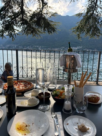 The image depicts an outdoor dining scene at a restaurant located in Pognana Lario, Italy, overlooking Lake Como. In the foreground, a table is set with plates, cutlery, glasses, a bottle of olive oil, a pizza, burrata cheese, a salad, breadsticks, and a bottle of wine chilling in an ice bucket. The background features the lake with a railing in front and a child seated on the left. Further in the distance, mountains rise beyond the lake, with a hazy sky above. The lighting suggests it is late afternoon or early evening, with the sun casting a warm glow on the scene. The overall ambiance is relaxed and convivial, with a focus on food and the picturesque natural surroundings.