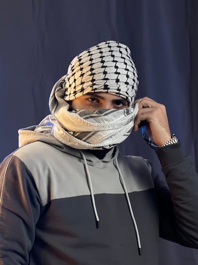 A person is wearing a black and white keffiyeh wrapped around their head and neck. Their eyes are visible, and they are looking towards the viewer. The person is wearing a two-toned hoodie, gray on one side and dark gray on the other. The sleeve of the hoodie is visible, and a silver watch is on the wrist. One hand is adjusting the keffiyeh. The background is a dark blue.