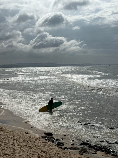A solitary individual stands in the shallow water at the edge of a beach, holding a colorful surfboard. The surfboard has a yellow and green design and is partially submerged in the water. The person, silhouetted against the bright, choppy sea, is wearing dark clothing. The ocean exhibits a high level of reflected light, creating a shimmering effect on its surface, with visible waves rolling towards the shore. The beach in the foreground consists of sand and scattered rocks. In the distance, a hazy silhouette of a coastline with mountains and wind turbines is visible beneath a heavily clouded sky. The overall atmosphere is overcast and possibly windy. The scene does not depict any specific landmarks or features that would definitively place it within Phan Thiết, Vietnam.