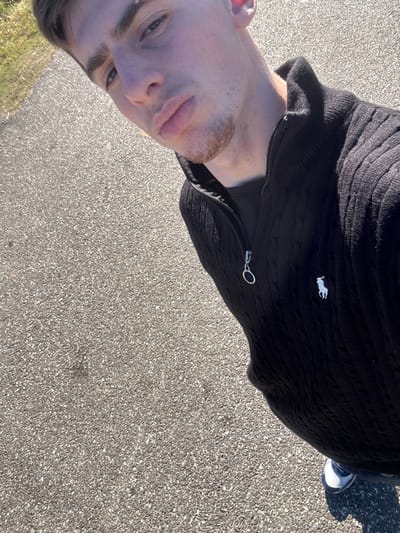 A man stands on asphalt. He wears a black zippered cable knit sweater with a white polo horse logo. He is angled, showing his face and part of his body. A foot in a sneaker is visible.
