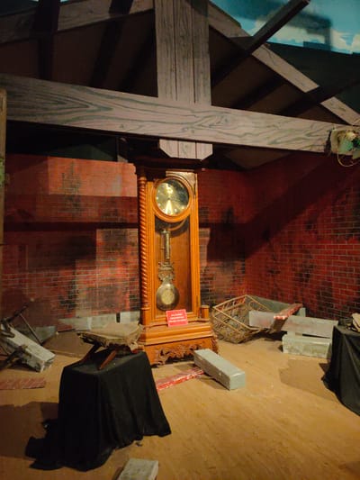 The image depicts a museum exhibit in Banda Aceh, Indonesia, featuring a grandfather clock standing against a red brick wall. The clock is made of wood and has a pendulum. In front of the clock, on the wooden floor, there is a dark cloth draped over a stand, with an old book placed on top of it. To the right of the clock, there is a display of what appear to be exhibit components, including a basket and some rectangular blocks. The room has a wooden ceiling with exposed beams, and a portion of a mural depicting a sky scene is visible at the top. The lighting suggests an indoor, possibly dimly lit, environment.