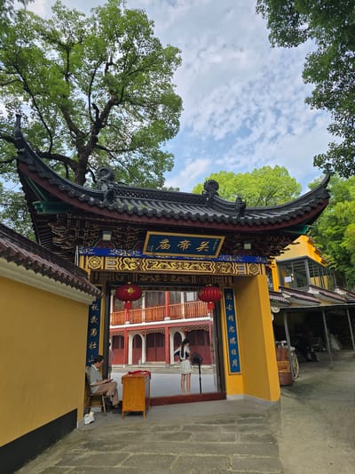 The image captures an entrance to a traditional Chinese courtyard, likely part of a temple or cultural site, situated in Xiaoshan District, China. The architectural style is characterized by a sweeping, ornate roof with dark tiles and intricate detailing, supported by colorful beams and columns. A prominent golden plaque with blue Chinese characters is affixed to the archway, above a large red lantern.
To the left of the entrance, a man in a grey shirt and beige pants sits on a wooden chair, looking at a mobile phone. Beside him is a wooden desk with a red cloth draped over it. To the right of the entrance, a young woman in a white dress stands near a pedestal fan, facing towards the courtyard.
The courtyard itself features a multi-story building with red balconies and windows, suggesting residential or administrative quarters within the complex. The ground is paved with stone, and the area is surrounded by lush green trees, indicating a natural or well-landscaped environment. The sky above is partly cloudy, with patches of blue visible. The overall impression is one of serenity and cultural heritage.