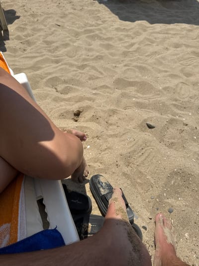 The image depicts a close-up view of two pairs of feet in the sand on a beach. One set of feet, belonging to a person sitting on a lounge chair, shows a tanned leg with sand dusting its surface. Beside these feet, a black sandal lies on the sand. The other pair of feet are in the foreground, covered in sand, with toes visible. Another black sandal is visible near these feet. The beach sand is light brown and appears textured with ripples. A shadow is cast from an unseen object, likely an umbrella or structure, across the sand in the upper portion of the image. In the background, faint outlines of beach furniture can be seen. The scene suggests a relaxed day at the beach, possibly in Sveti Vlas, Bulgaria, known for its sandy shores and resort atmosphere.