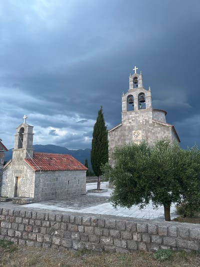 The image shows a view of a small stone church with a bell tower, partially obscured by a large olive tree. The church has a tiled red roof and a light gray stone facade. The bell tower is also made of stone and features several bells. In the foreground, a stone wall runs along a pathway paved with light-colored stones. In the background, dark, dramatic clouds suggest impending rain or a storm. Mountains are visible in the distance, and a tall, dark green conifer tree stands behind the church. The overall scene is one of solemnity and perhaps a touch of drama due to the weather. The lighting suggests it might be late afternoon or early evening, given the dramatic sky. No people or text are visible in the image. The location is Kovačko Polje, Montenegro.